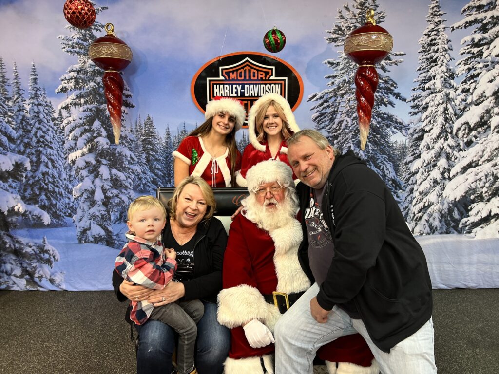 Santa sits with a family of three at Gowanda Harley-Davidson. His two elves stand behind him as he sits on his throne with and adult man (Grandpa) sitting on his lap and the Grandmother and child sitting next to himi. They are pictured against the backdrop of a Harley-Davidson logo and a wintry scene of snow covered evergreen trees. Everybody is smiling and happy and looking forward to celebrating Christmas 2022. 