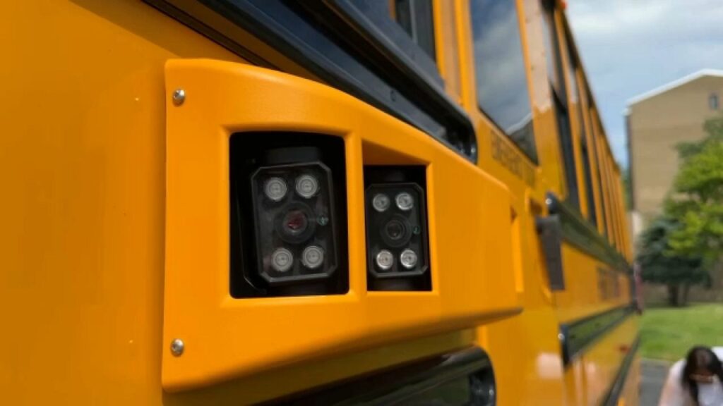 A camera mounted on the side of a yellow school bus that is designed to capture images of drivers that illegally pass while the bus's lights are flashing and it is unloading or loading students.